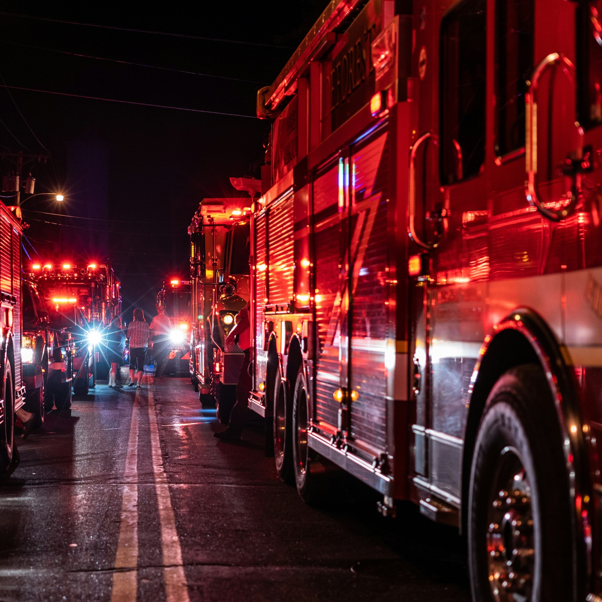 Multiple fire engines with flashing lights lined along a roadway at night, with responders and people visible between the vehicles. Multiple fire engines with flashing lights lined along a roadway at night, with responders and people visible between the vehicles.