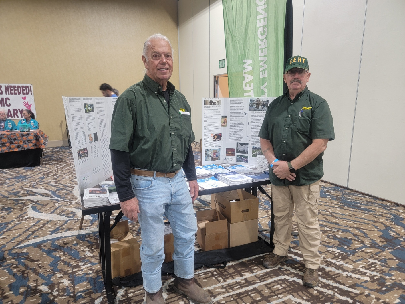 Two CERT volunteers wearing green shirts and a CERT cap stand behind an information table with brochures and display boards at a community event. A green CERT banner is visible behind them, and other booths and attendees can be seen in the background.
