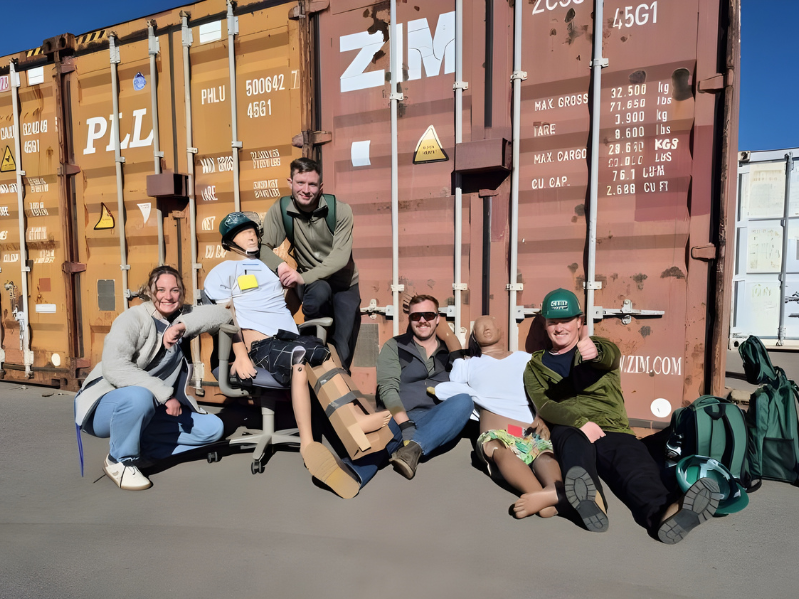 CERT team members pose outside in front of shipping containers with training mannequins, wearing green helmets and safety gear. The group smiles and gives thumbs up while sitting and kneeling beside the mannequins used for emergency response training.