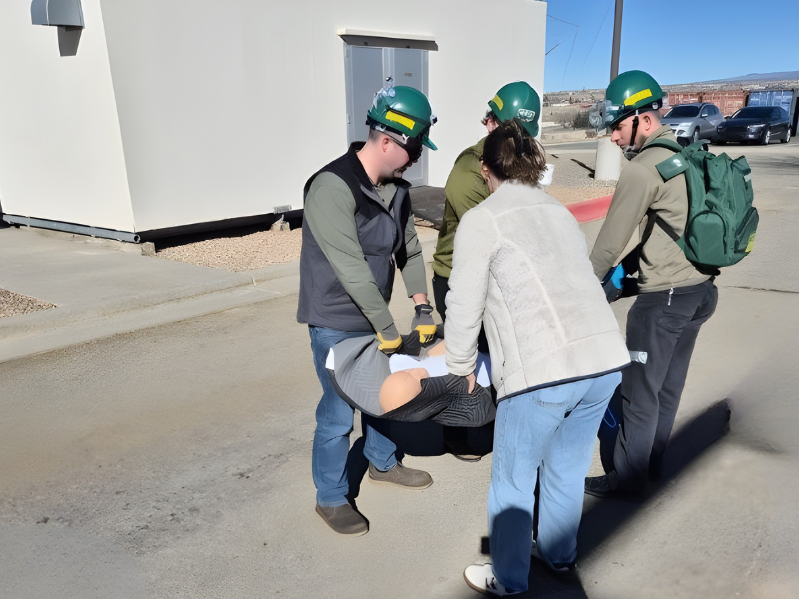 CERT team members wearing green helmets and safety gloves carry a training participant on a stretcher outside a building during a hands-on emergency response exercise.
